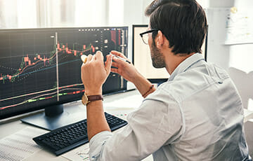 Successful Trading. Back View Of Young Bearded Trader In Formal Wear Pointing On The Data On Computer Screen With Pen And Holding Bitcoin In One Hand While Working His Modern Office.
